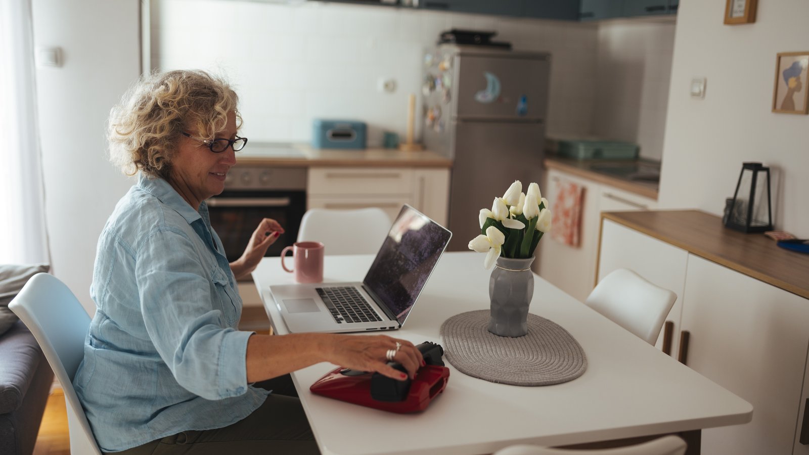 Femme âgée souriante assise à la table de sa cuisine, devant un ordinateur portable, une tasse et un vase de tulipes blanches