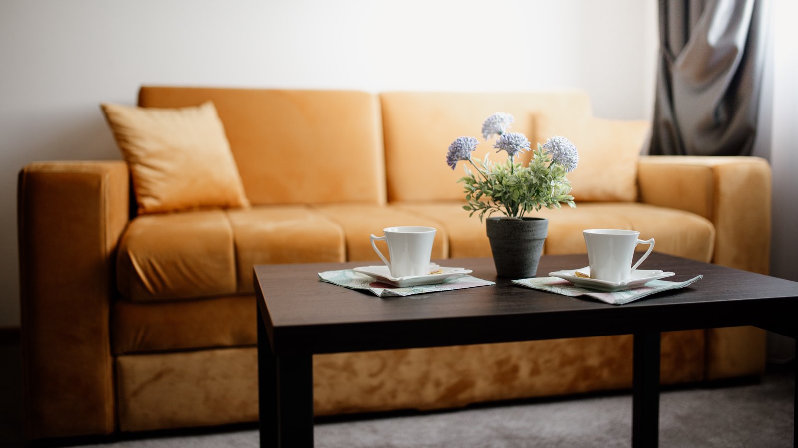 Warm, contemporary living room: ochre velvet sofa, matching cushions, dark wooden coffee table with two cups and a bunch of flowers