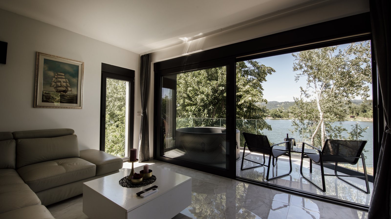 Bright living room with a pale corner sofa, opened up by a large sliding picture window onto a terrace, trees and a lake