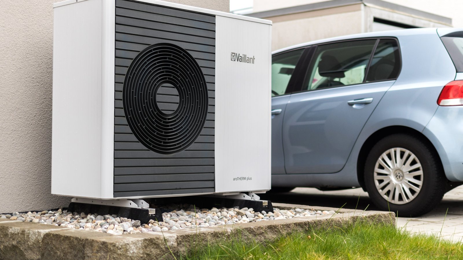 Outdoor unit of an air-to-water heat pump standing on a bed of gravel against the front wall of a house, with a car parked alongside