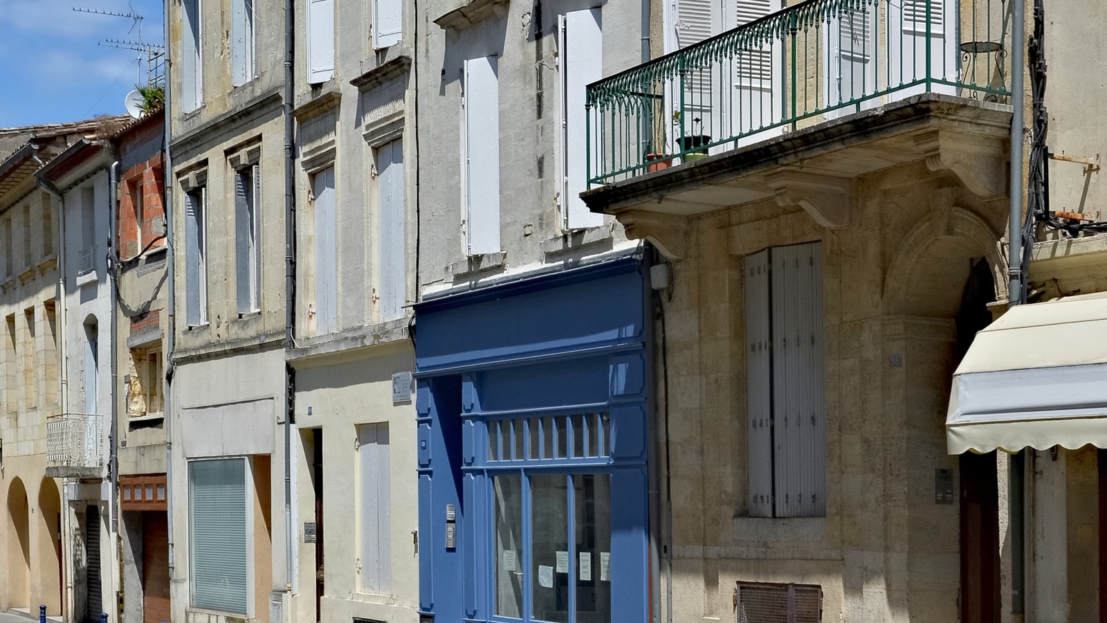 Street in central Libourne lined with terraced limestone townhouses, white shutters, a wrought-iron balcony and a blue shopfront