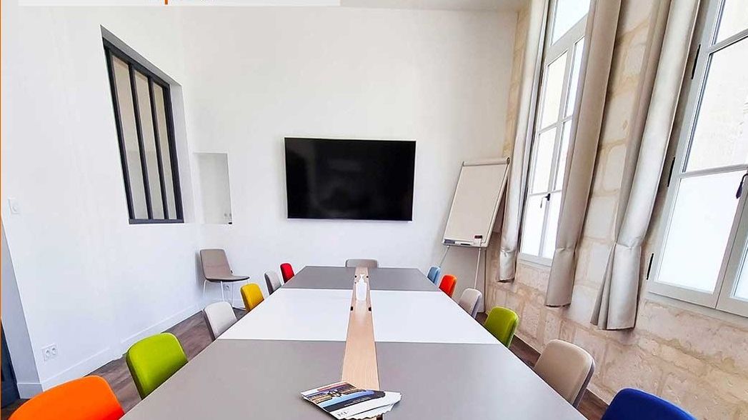 Salle de réunion rénovée avec grande table blanche, chaises colorées et écran