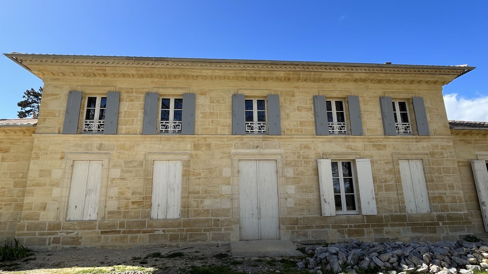 Façade en pierre de taille restaurée avec volets gris, sous un ciel bleu