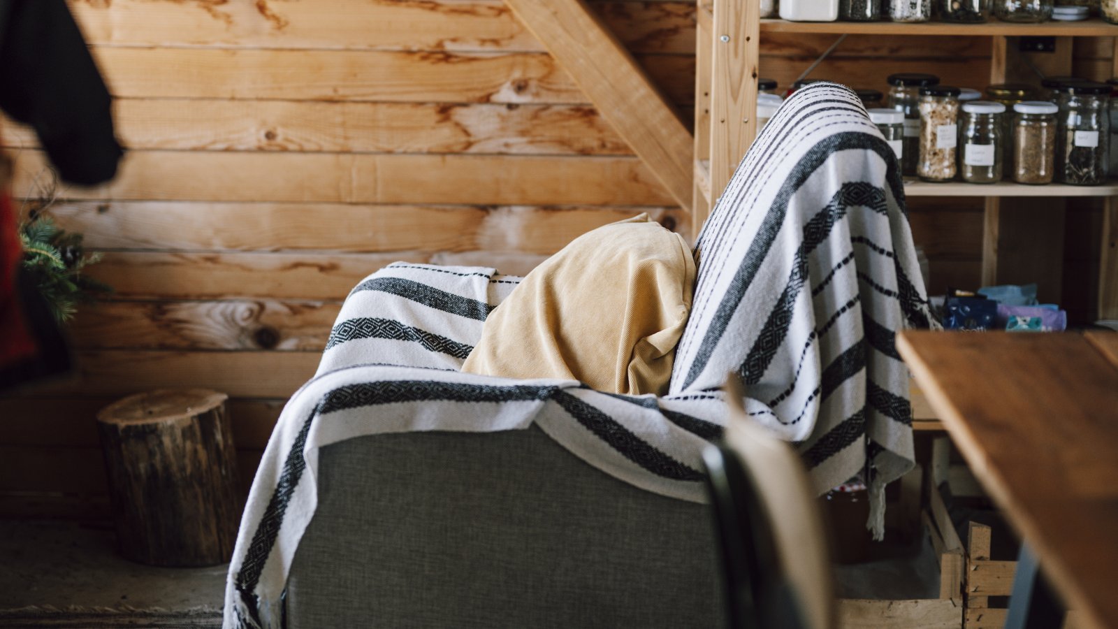 Armchair covered with a striped throw and a mustard cushion in front of a wall of raw wooden planks, with a log and a wooden shelf holding jars