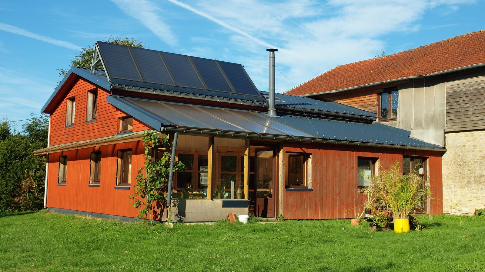 Maison basse consommation en bardage bois rouge, panneaux solaires sur le toit, véranda vitrée et jardin, sous un ciel bleu